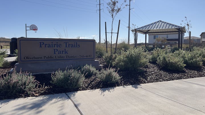 A park sign reading "Prairie Trails Park," a gazebo, basketball hoop, plants, and power lines against a bright sky.