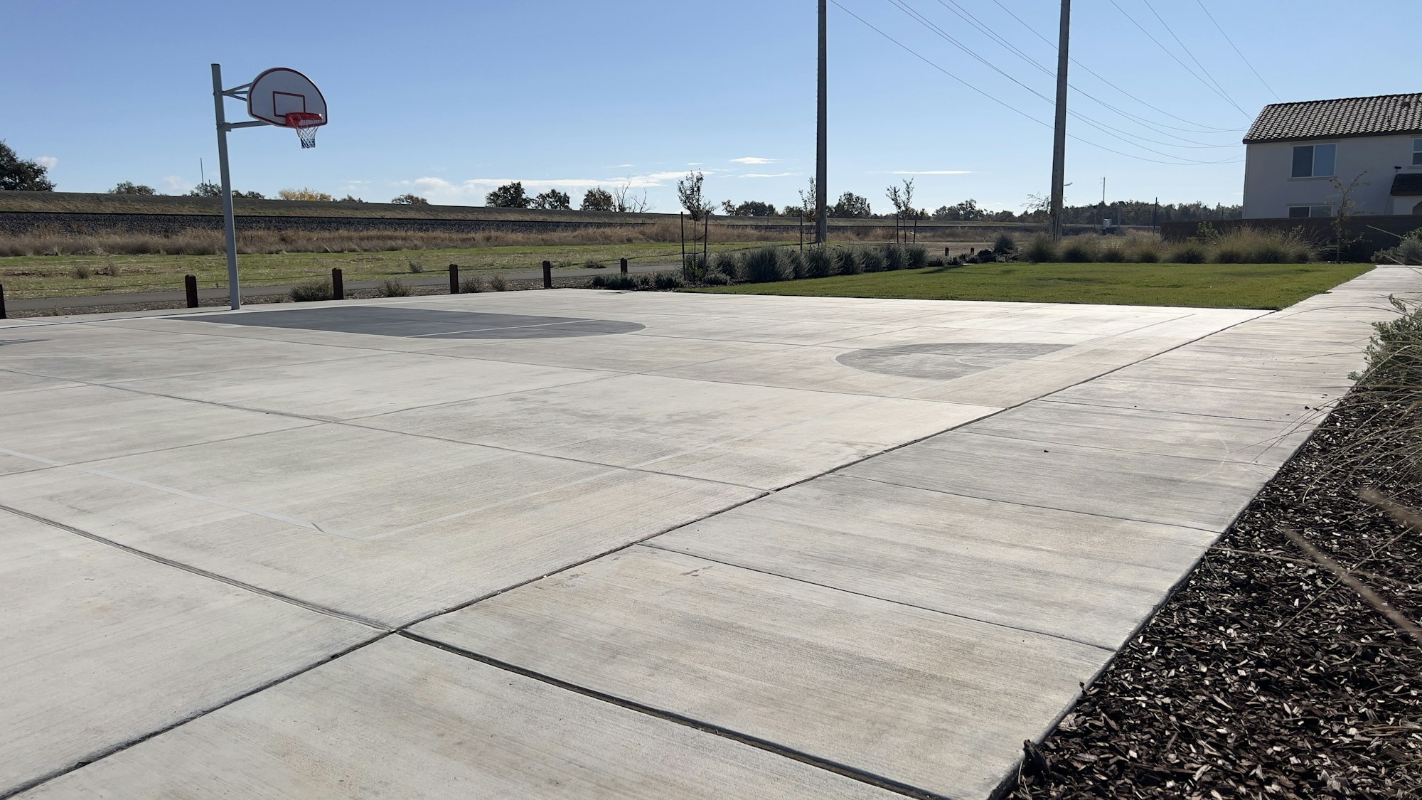 A basketball court with a hoop, concrete pavement, nearby grass, and a house in the background.