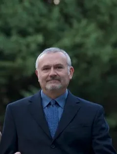 A person in a suit stands outdoors in front of green foliage, appearing confident and poised.