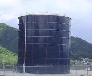 The image shows a large cylindrical storage tank, likely for water or fuel, situated near green hills.