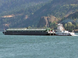 A tugboat is pushing a large barge on a river with forested hills in the background.