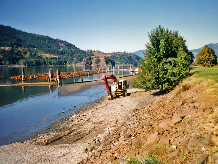 Excavator on a rocky shore by a river with floating logs, surrounded by hills and trees.