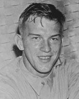 A black and white photo of a smiling man in front of a brick wall background.