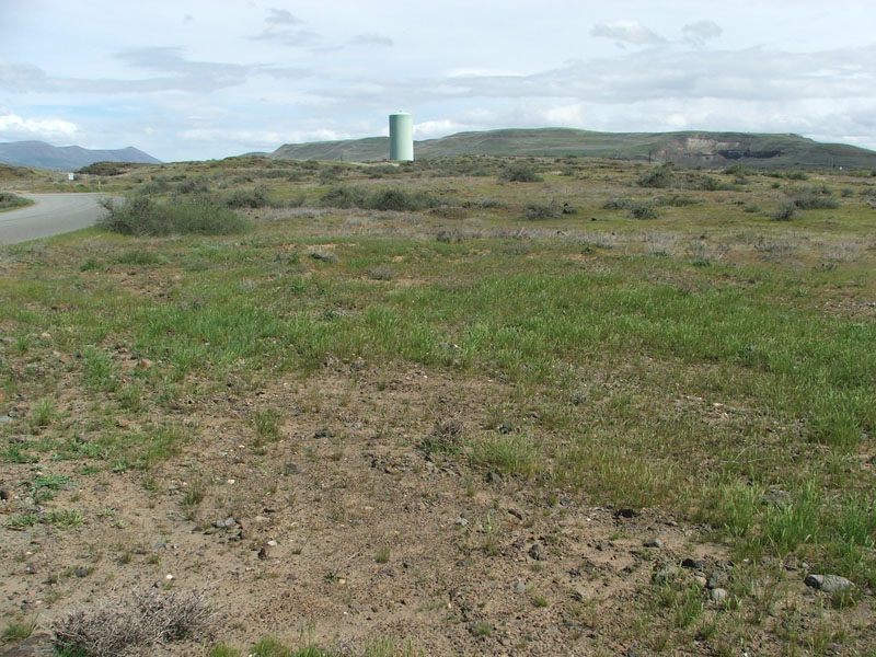 A barren landscape with sparse vegetation, a road, and a tall cylindrical structure in the distance.