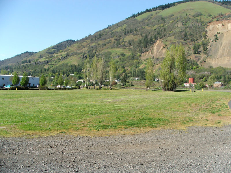 Open grassy field with trees, buildings, and a hillside in the background under a clear sky.