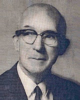 An elderly person wearing glasses, in a suit and tie, looking slightly to the side. The image is in black and white.