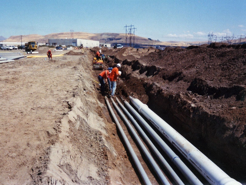 Workers in a trench installing large pipes at a construction site with vehicles and equipment in the background.