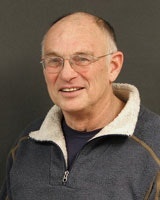 A smiling older man with glasses and short hair, wearing a sweater with a collar, set against a dark background.