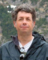 A person wearing a jacket stands outdoors with trees and mountains in the background.