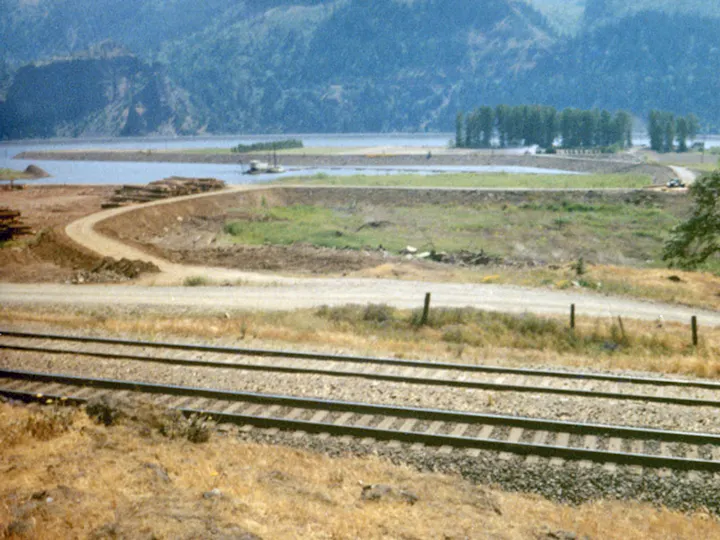 Railroad tracks in the foreground, a dirt road, grassy fields, a river, and forested hills in the distance.