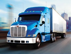 A blue semi-truck with a trailer drives on a road, with a blurred cityscape in the background.