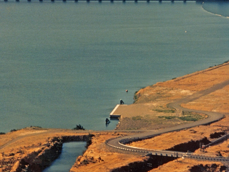 A curving road by a body of water with a small dock, surrounded by dry, brown terrain.