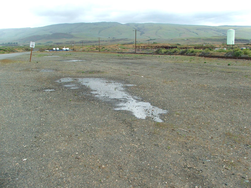 Gravel road with puddles, speed limit sign, hills in background, railroad tracks, and a water tower.