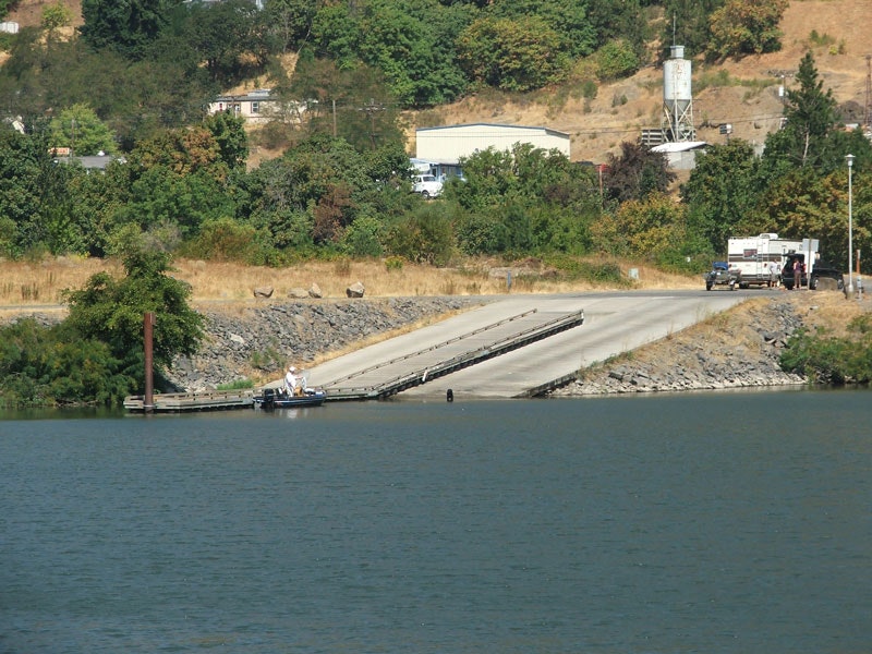 A boat ramp leads into a calm body of water with a small boat docked. Vehicles and trees are in the background.