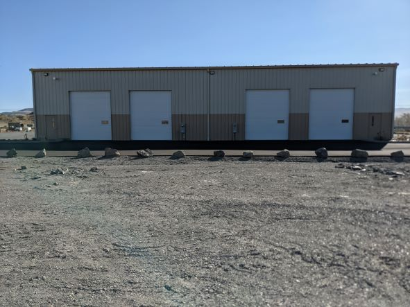 A large warehouse building with three closed garage doors, surrounded by a gravel lot.