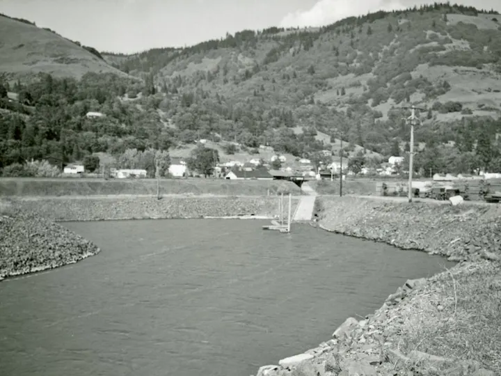 River with rocky banks, small town in the background, surrounded by hills and trees.