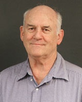 Older man in a light gray shirt, photographed against a dark background.