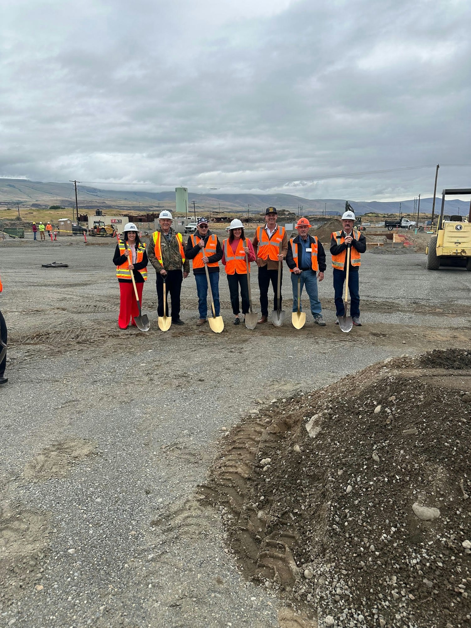A group of people in hard hats and orange vests stand on a construction site holding shovels.