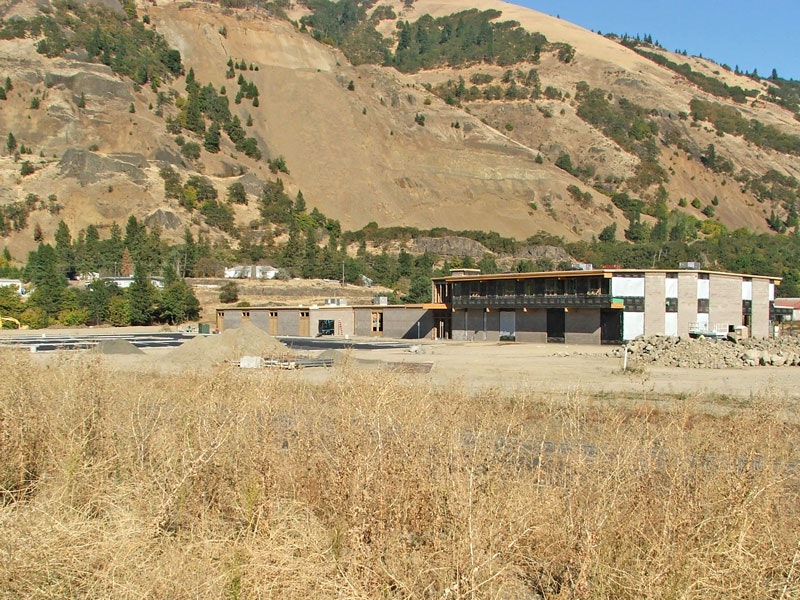 Construction site with buildings in progress, surrounded by dry grass and situated near a hillside with trees.