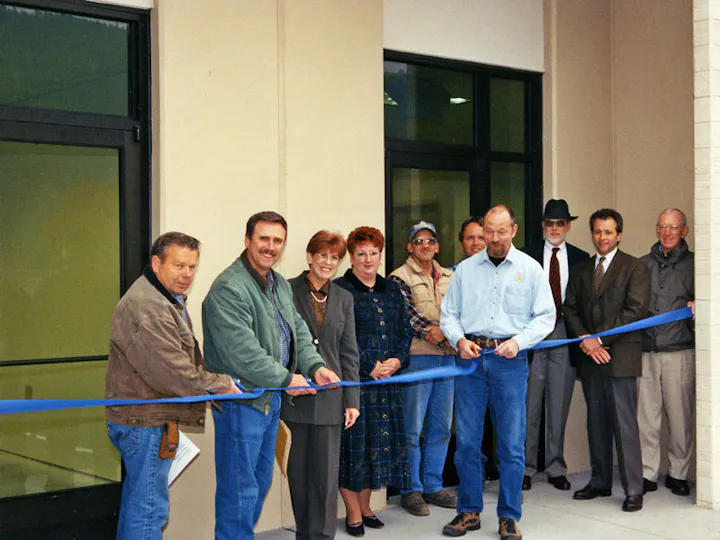 A group of people at a ribbon-cutting ceremony, holding a blue ribbon outside a building.