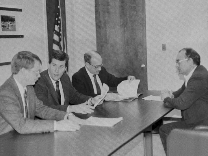 Four men in suits sitting at a table, discussing documents in an office setting. An American flag is visible in the background.