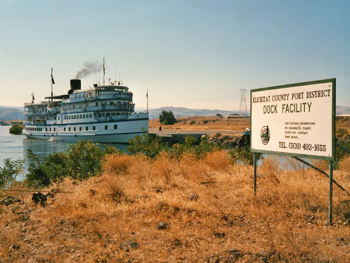 A large ship at Klickitat County Port District dock facility beside a dry, grassy area with a sign in the foreground.