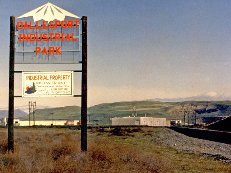 Sign for Dallesport Industrial Park; offers industrial property for lease or sale. Background shows industrial buildings and landscape.