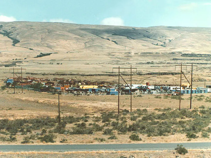 A desert landscape with hills, sparse vegetation, power lines, and a small settlement of colorful buildings and structures.