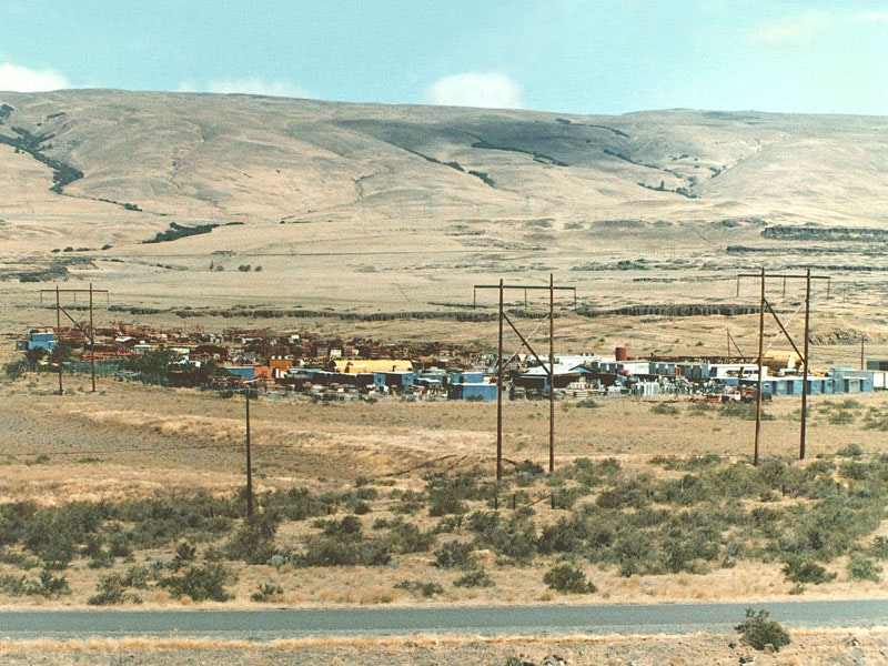 A desert landscape with hills, sparse vegetation, power lines, and a small settlement of colorful buildings and structures.