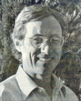 A black and white image of a person wearing glasses and a collared shirt, smiling outdoors.