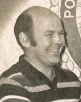 A smiling person in a striped shirt, standing against a backdrop with partially visible text.