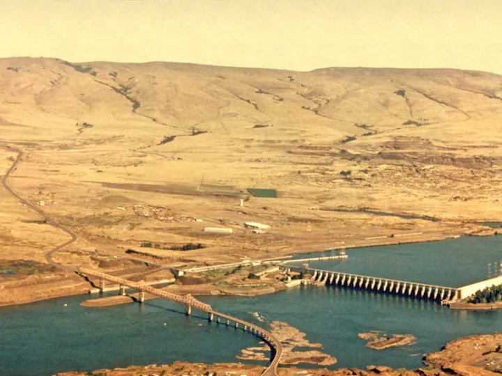 A dam and bridge span a river with arid hills and a sparse landscape in the background.