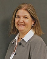 A person with shoulder-length hair, wearing a gray blazer and white shirt, smiling against a gray background.