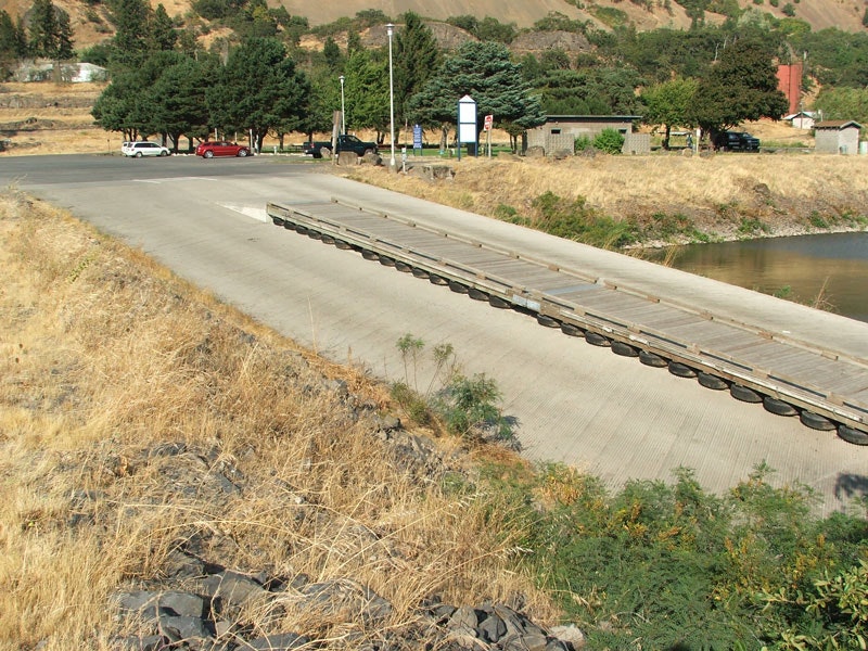 A concrete boat ramp leading into a water body, surrounded by dry grass, trees, and parked cars in the distance.