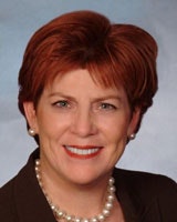 A person with short red hair and a pearl necklace, wearing a dark jacket, smiling against a neutral background.