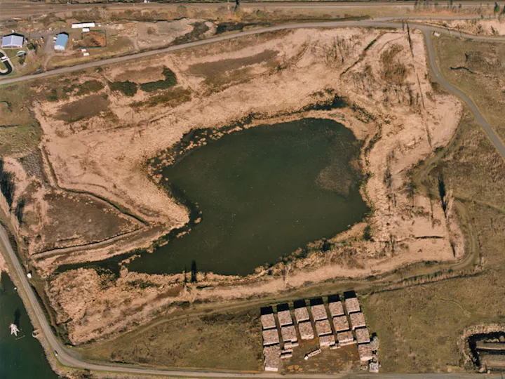 Aerial view of a pond surrounded by vegetation and dirt roads, with several rectangular structures in the foreground.