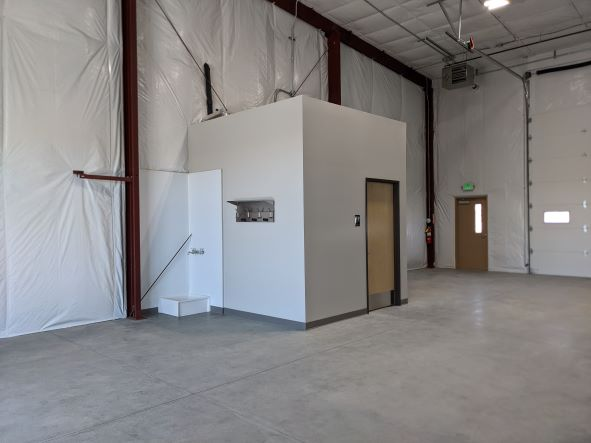 A small room with doors inside a large warehouse-like space with white walls and exposed beams.