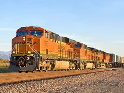 A freight train with orange locomotives travels along a gravel track under a clear sky.