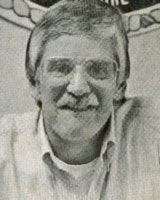 A man with glasses, mustache, and a light-colored shirt smiling at the camera.