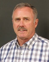 A man in a checkered shirt is standing against a plain background.