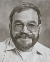 A bearded man is wearing glasses and a collared shirt, smiling in a grayscale portrait photo.