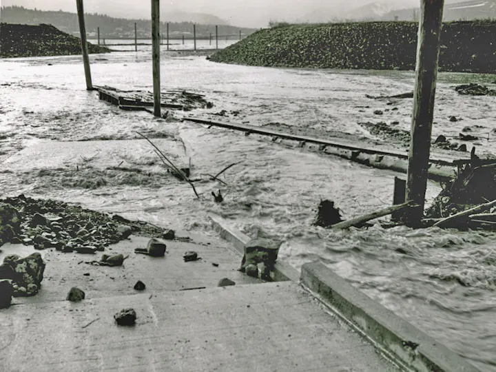 Flooded area with fast-flowing water, debris, and damaged infrastructure.