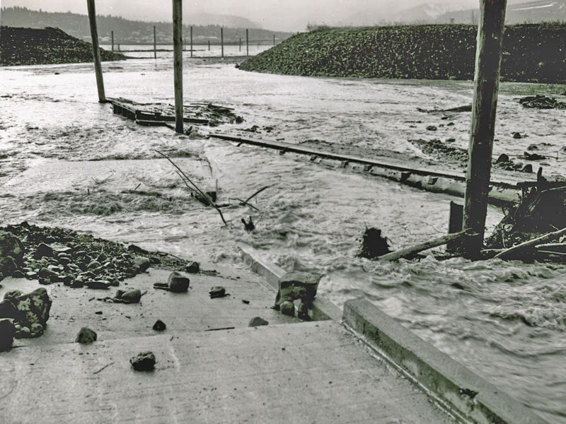 Flooded area with fast-flowing water, debris, and damaged infrastructure.