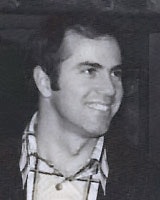 A person smiling, wearing a checkered shirt and a jacket, in a black and white photo.
