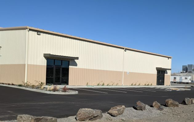 A beige and tan industrial building with large windows and an empty parking lot, featuring rocks in the foreground under a blue sky.