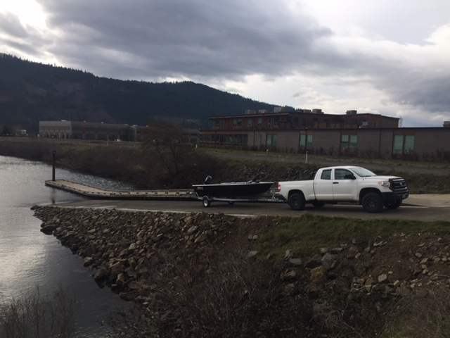 A white pickup truck towing a boat on a trailer near a ramp by a body of water, with buildings and hills in the background.