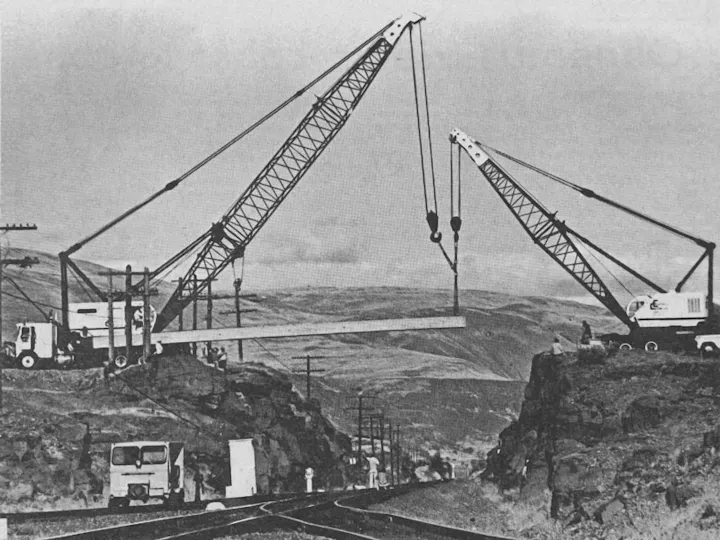 Two cranes lifting a horizontal beam over a valley with hills in the background.
