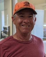 A person smiling, wearing an orange cap and a red shirt, standing indoors near a window.