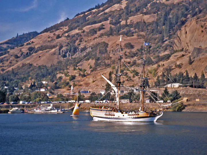 A sailing ship on a river with hills in the background, accompanied by smaller boats.