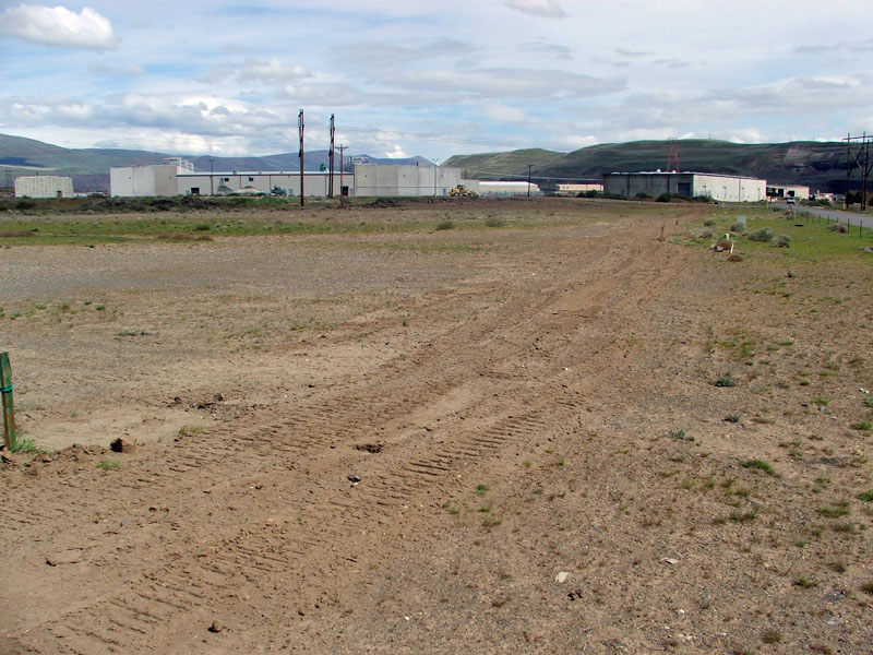 Industrial buildings near a dirt field with some vegetation and distant hills under a partly cloudy sky.